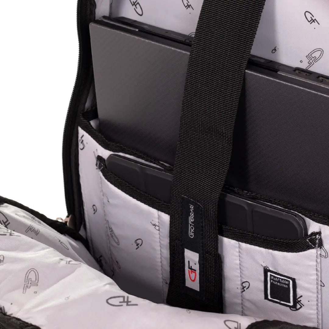 Close-up of a laptop backpack interior showing a black laptop secured in a padded compartment, with a black webbing strap and a small brand patch on the fabric.