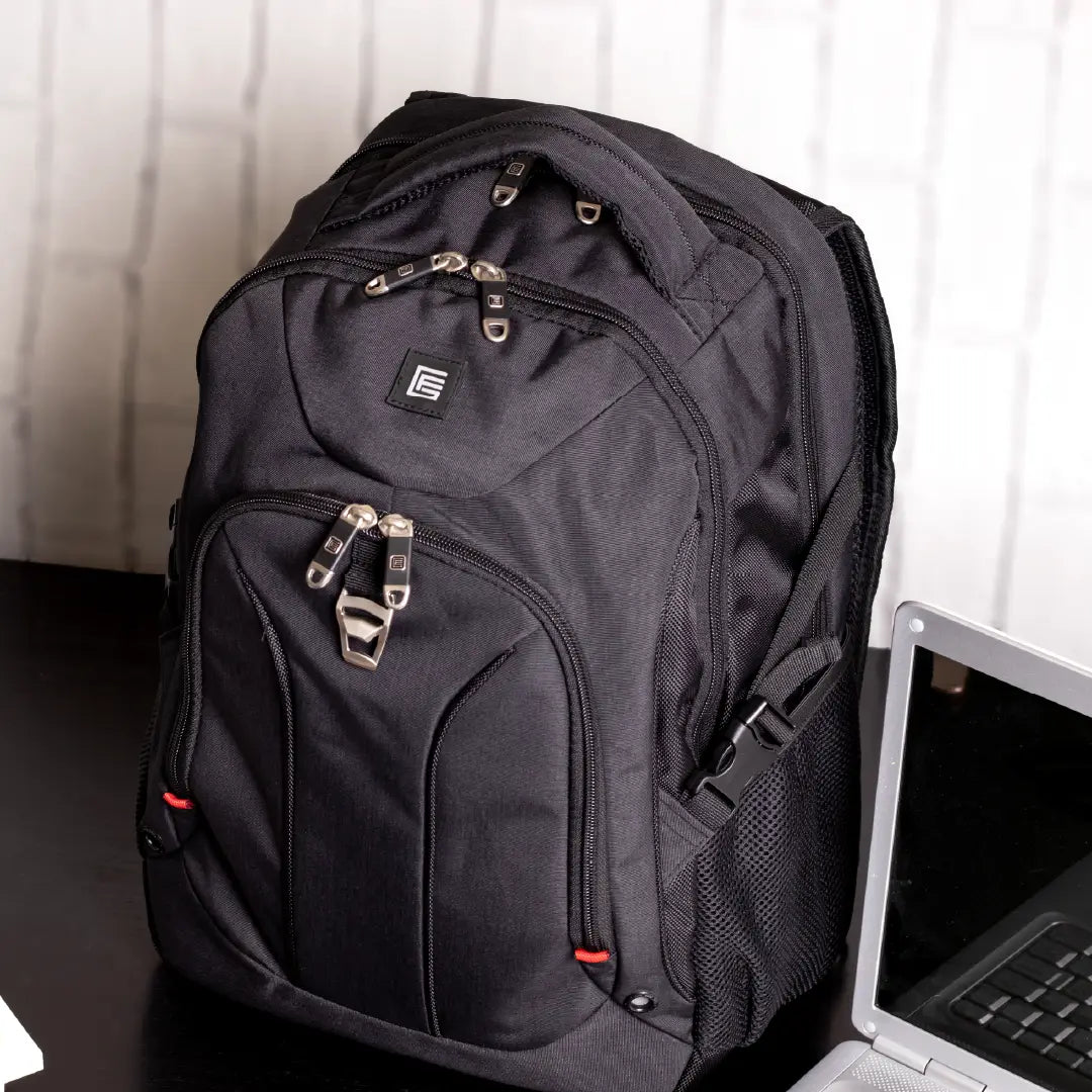 A black laptop backpack with multiple zippered compartments and a side mesh pocket, shown on a dark tabletop next to an open laptop in front of a light-colored paneled wall.