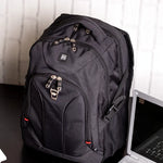 A black laptop backpack with multiple zippered compartments and a side mesh pocket, shown on a dark tabletop next to an open laptop in front of a light-colored paneled wall.
