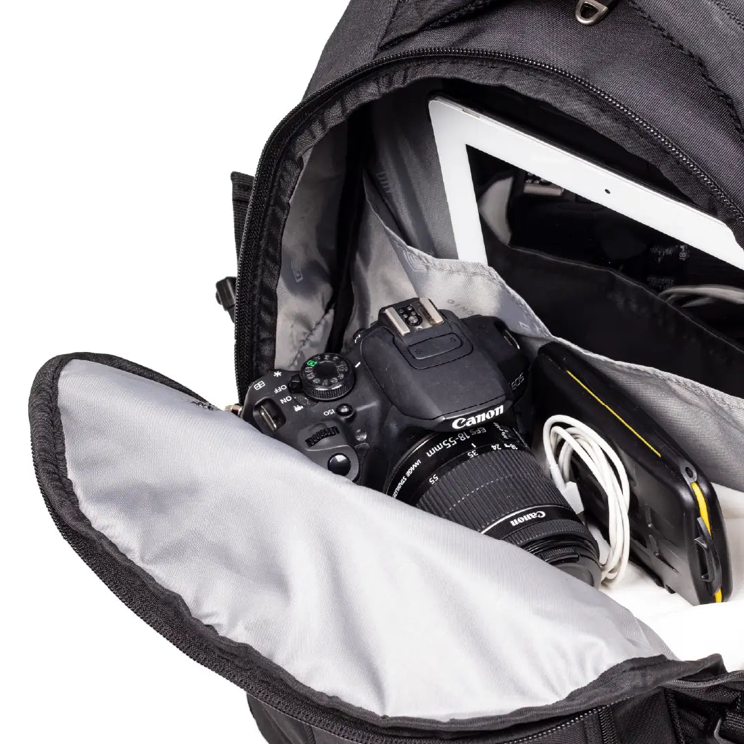 Open black camera backpack showing interior compartments with a Canon DSLR camera, tablet, and accessories inside on a white background.