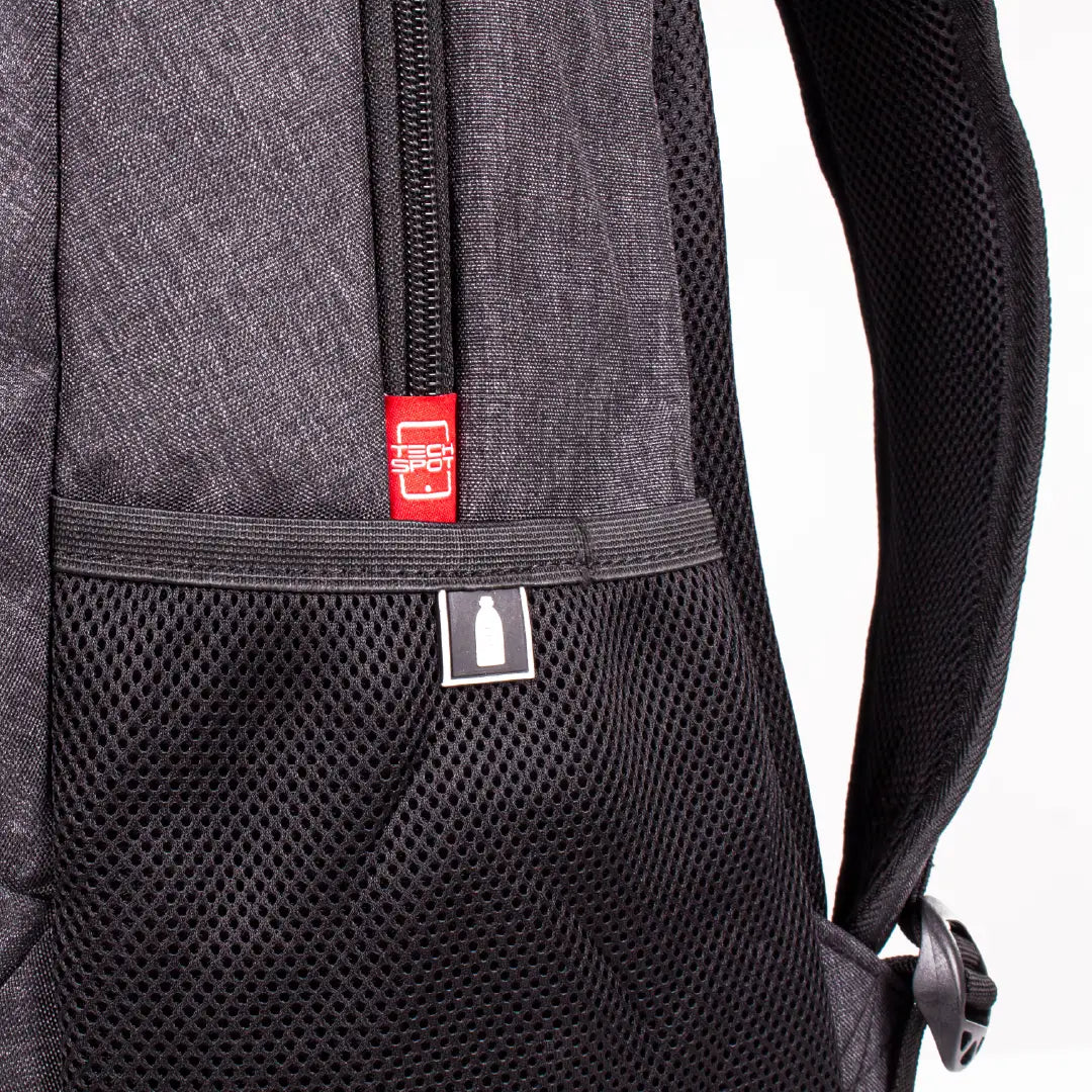Close-up of a black and dark gray backpack showing the front mesh pocket, a black zipper, and a small red brand tag with a white bottle icon.