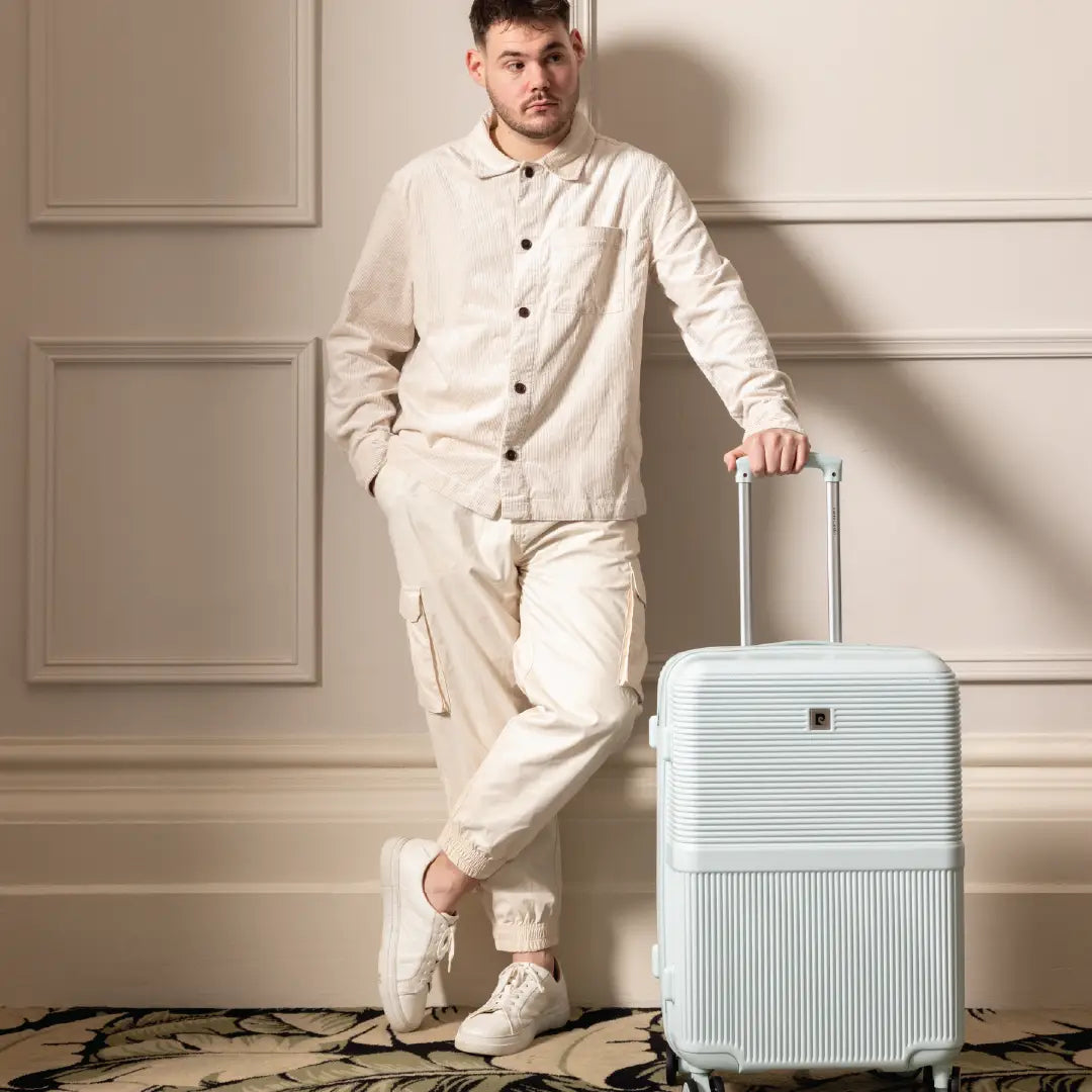 Man in a white outfit holding a light blue suitcase against a neutral wall.