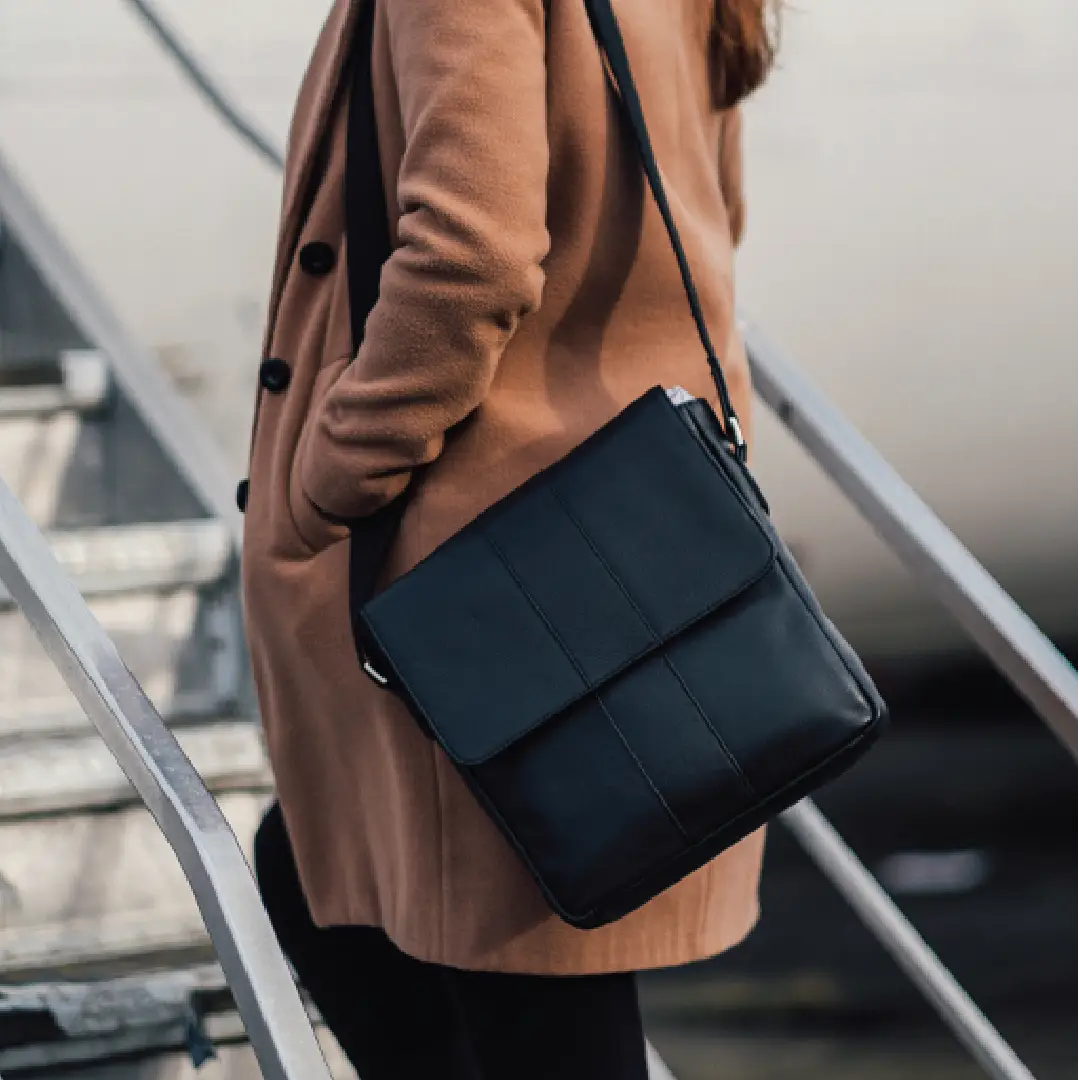 A person wearing a camel coat carries a black leather crossbody bag with a structured flap and shoulder strap while standing on metal stairs.