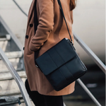 A person wearing a camel coat carries a black leather crossbody bag with a structured flap and shoulder strap while standing on metal stairs.