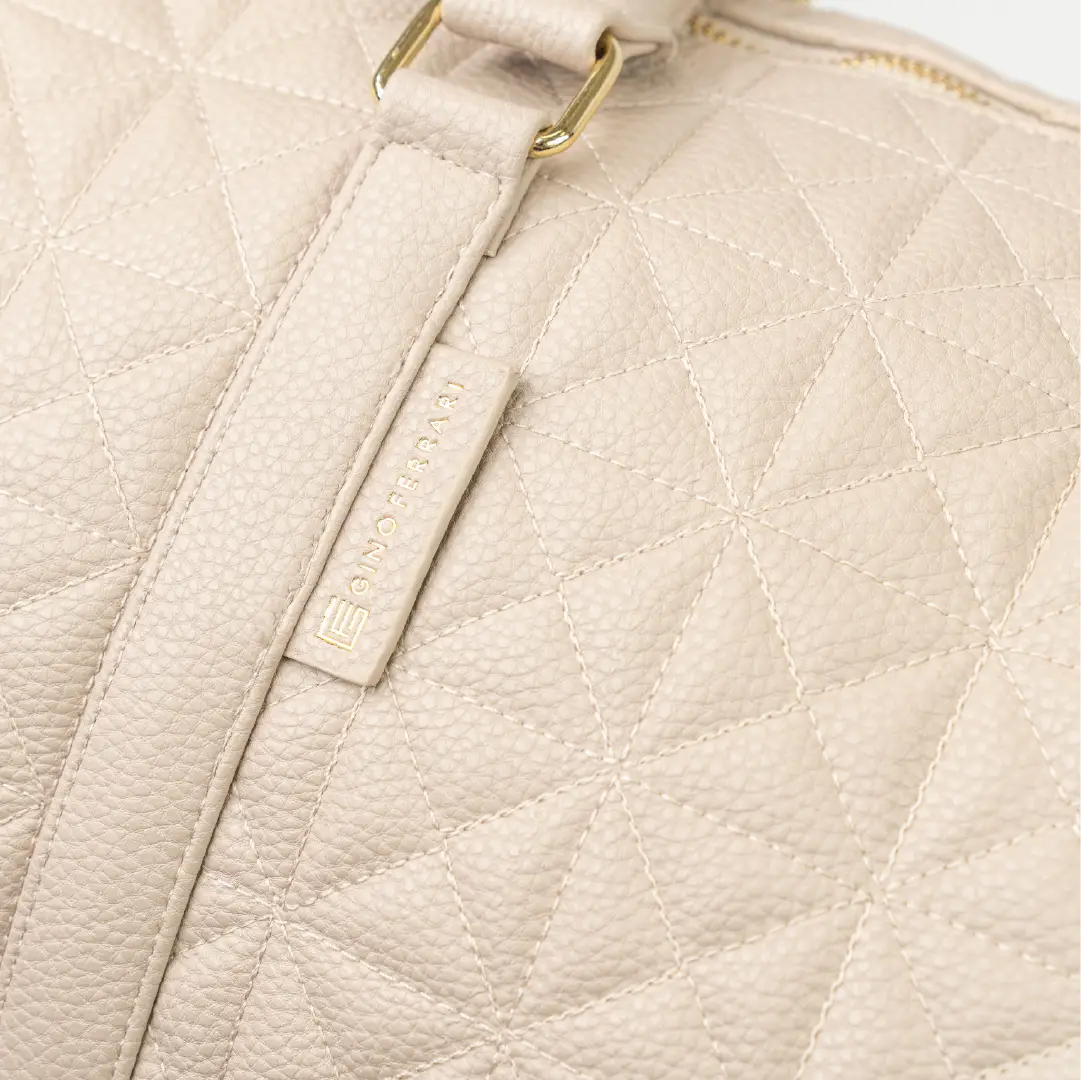 A close-up of a beige bag with a brand logo, patterned stitching and a gold zip.