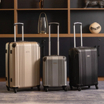 Three suitcases of different colors on a wooden floor with a dark wall and shelves in the background.