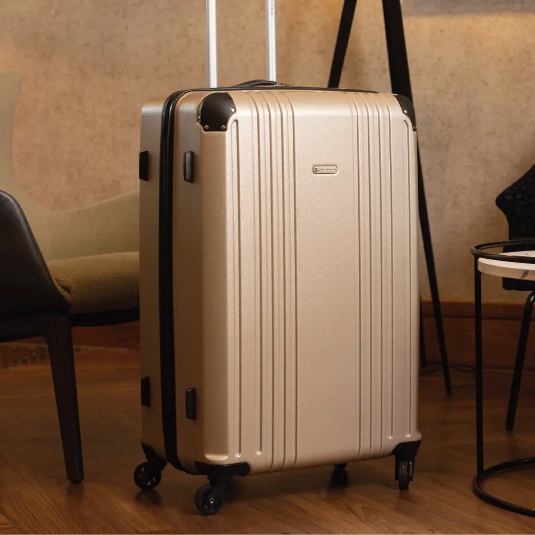 Gold hard-shell suitcase with a visible brand logo and black accents in a hotel reception setting.