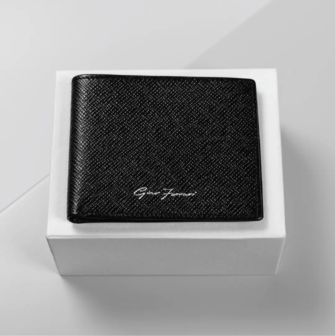 A black textured leather bifold wallet with a silver-tone 'Gino Ferrari' signature logo, presented on a white square gift box against a light gray background.