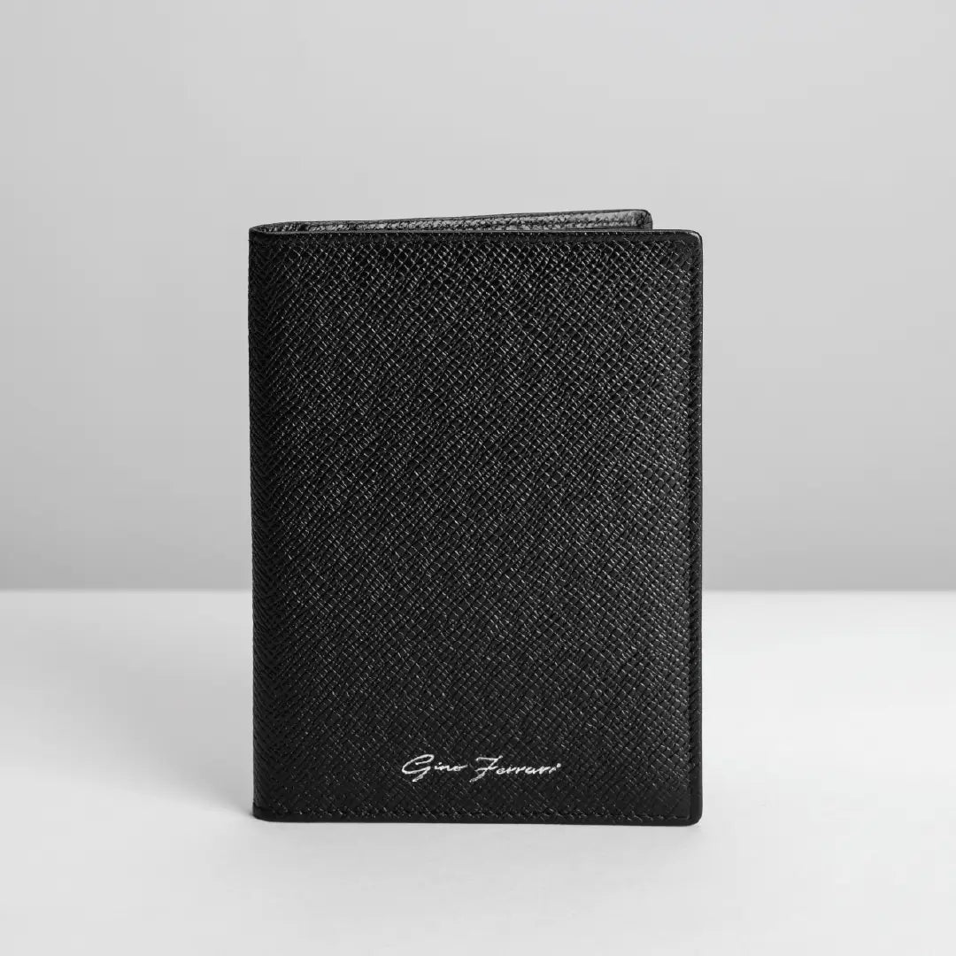 A black textured leather passport cover with a silver-tone 'Gino Ferrari' signature logo, standing upright on a light grey surface against a neutral grey background.
