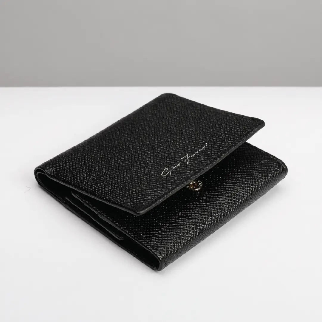 A black textured leather bifold wallet with a small gold-tone snap button and a silver-tone script logo on the front, displayed on a clean white surface with a gray background.