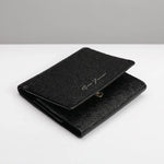 A black textured leather bifold wallet with a small gold-tone snap button and a silver-tone script logo on the front, displayed on a clean white surface with a gray background.
