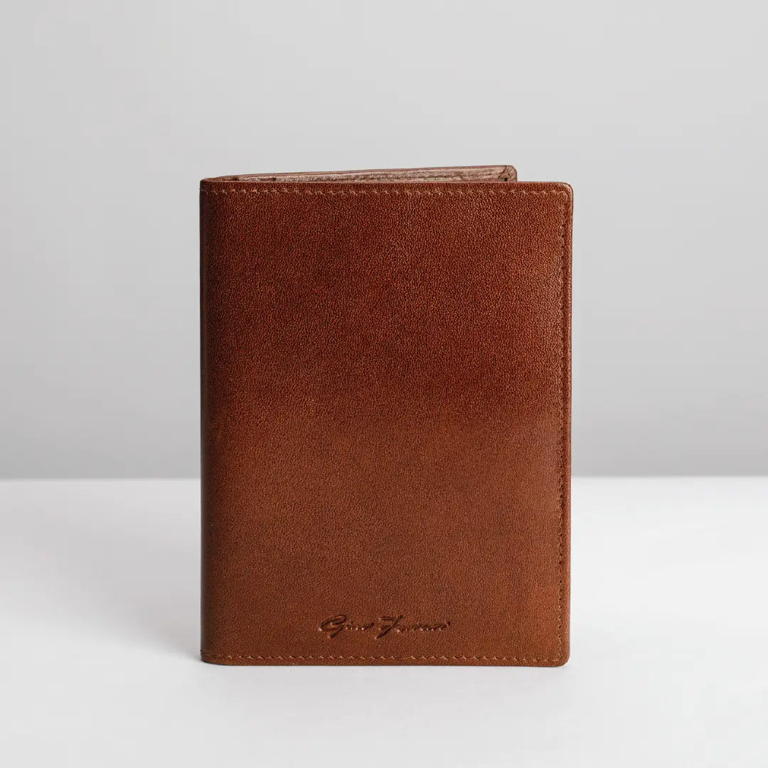 A brown leather passport cover or wallet standing upright on a white surface against a light gray background. A small debossed logo is visible near the bottom.