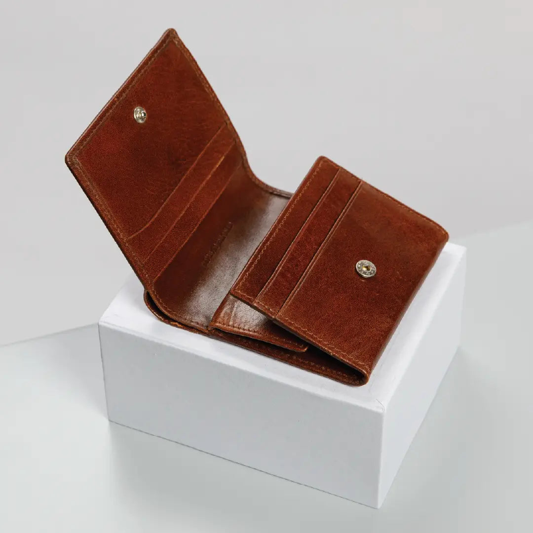 A brown leather bi-fold wallet shown open, resting on a white display block against a light gray studio background.
